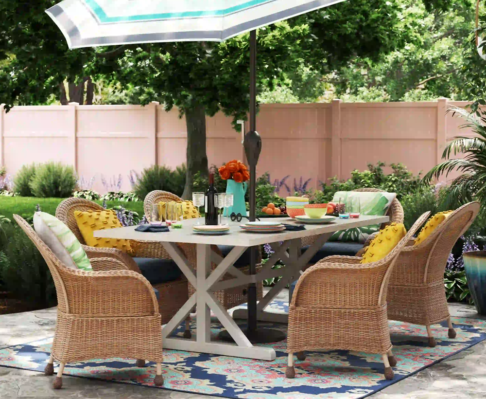 Photorealistic 3D rendering of an outdoor patio furniture setup with wicker chairs, a wooden table, colorful pillows, and a garden umbrella