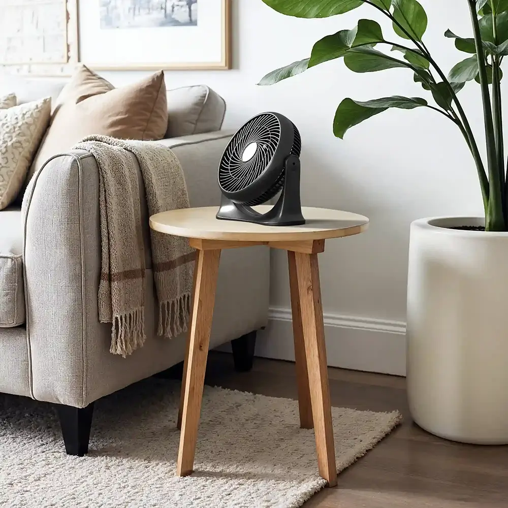 A small black table fan placed on a wooden side table in a modern living room with a beige couch and a large plant.